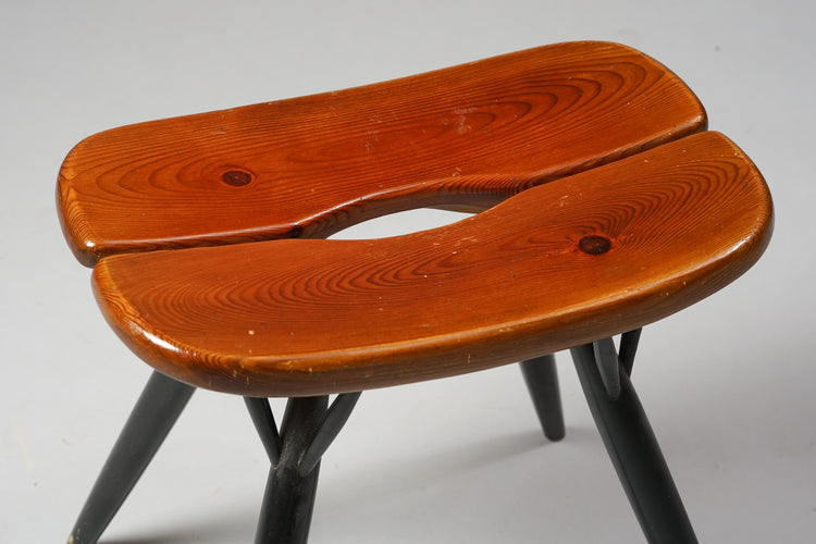 Wooden stool with a curved seat. The wood colored seat has a slit in the middle and the legs are painted balck.