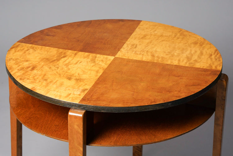 Round table made of birch and plywood. The tabletop has two colors, lighter and darker brown. There is a shelf under the top.