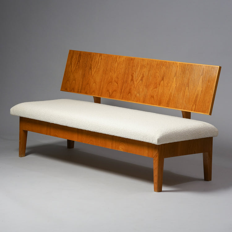 Puusta tehty sohva, jossa valkoinen kankaasta tehty istuin. Sohvan selkänoja on myös tehty puusta.